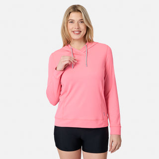 A smiling woman with long blonde hair wears the Free Country Women's SunFree UPF Hoodie in pink and black shorts, standing against a plain light gray background.