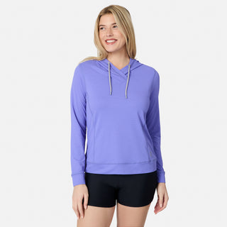 A woman smiles against a white background wearing the Free Country Women's SunFree UPF Hoodie in light purple and black shorts. The hoodie features moisture-wicking fabric and UPF 50+ sun protection.
