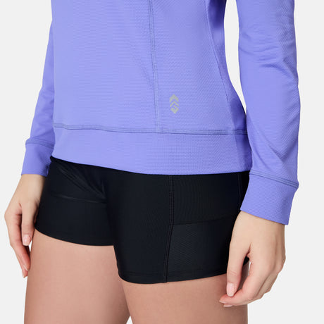 A person wears the Free Country Women's SunFree UPF Hoodie, a long-sleeve light purple athletic top with moisture-wicking fabric, and black shorts. Only the torso and upper legs are visible against a plain white background.