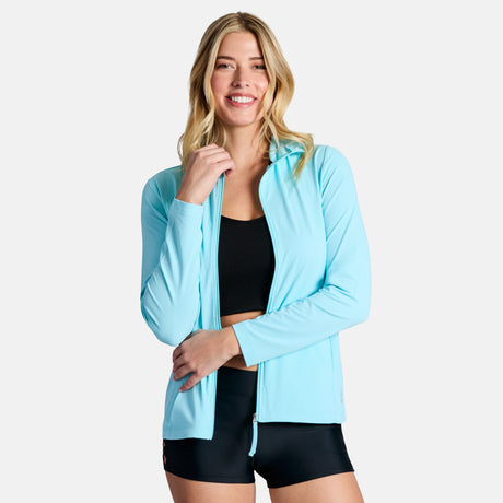 A smiling woman with long blonde hair wears the Free Country Women's SunFree Full Zip UPF Sunshirt in light blue over a black sports bra and black shorts, standing against a plain light background.