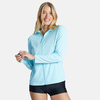 A smiling woman with long blonde hair wears the Free Country Women's SunFree Full Zip UPF Sunshirt in light blue and black shorts, standing against a plain white background.