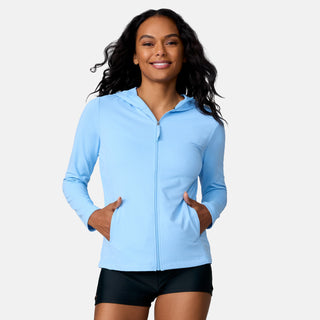 A woman with long, wavy hair smiles while wearing the Free Country Women's SunFree Full Zip UPF Sunshirt in light blue and black shorts, standing with her hands in her pockets against a plain white background.