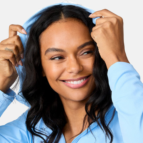 A smiling woman with long dark hair pulls up the hood of her Free Country Women's SunFree Full Zip UPF Sunshirt, a moisture-wicking cover-up that keeps her cool and comfortable as she looks to the side.