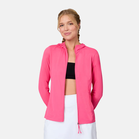 A woman with light hair in a low ponytail smiles gently, wearing the Free Country Women's SunFree Full Zip UPF Sunshirt over a black crop top and white skirt, standing against a plain background with her hands behind her back.