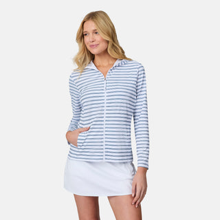 A woman wears the Free Country Women's SunFree Full Zip UPF Sunshirt in light blue and white stripes with a white skirt, standing with one hand in her pocket and smiling slightly against a plain white background.