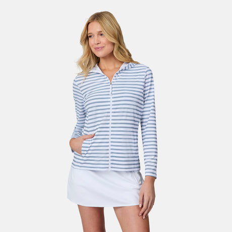 A woman wears the Free Country Women's SunFree Full Zip UPF Sunshirt in light blue and white stripes with a white skirt, standing with one hand in her pocket and smiling slightly against a plain white background.