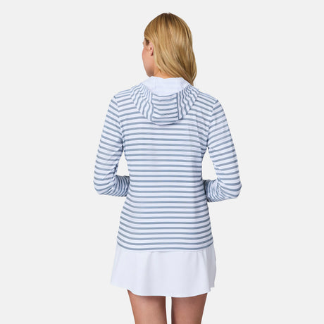 A woman with blonde hair stands facing away, wearing the Free Country Women's SunFree Full Zip UPF Sunshirt in light blue and white stripes over a white skirt. The sunshirt features a hood and is made from recycled fabric against a plain white background.