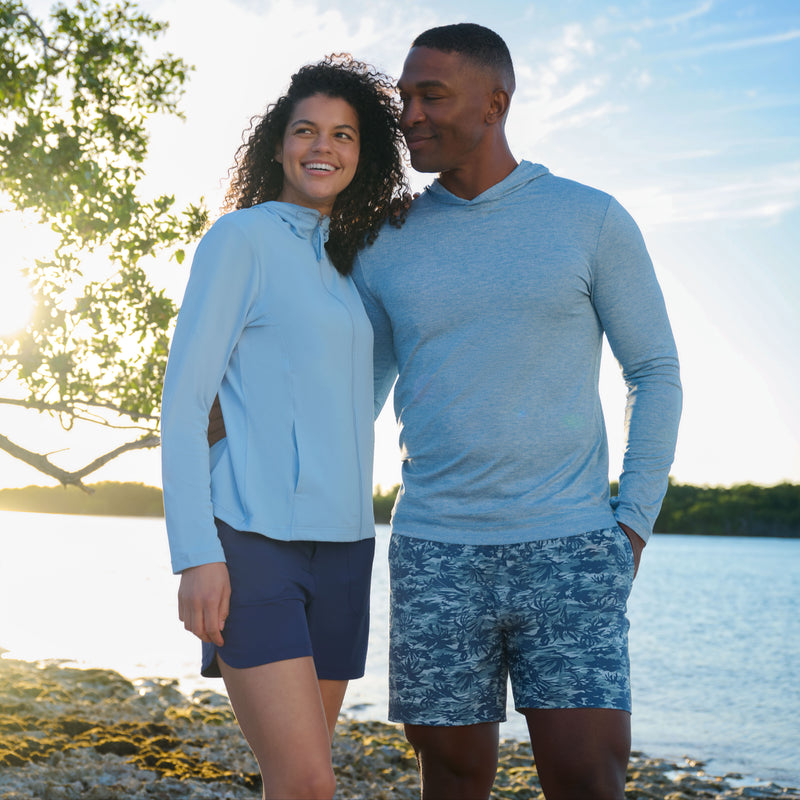 A smiling couple stands on a rocky shoreline at sunset. The woman wears the Free Country Women's SunFree Full Zip UPF Sunshirt, while the man sports a gray hoodie and blue patterned shorts. Trees frame the scene under a bright sky.
