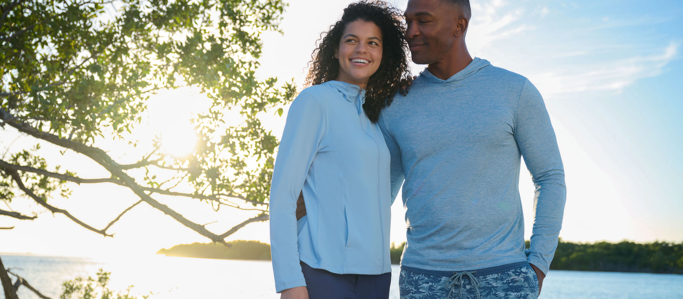 Man and woman in matching blue long-sleeve shirts standing by a body of water with trees in the background.