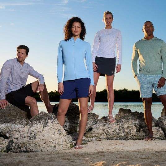 Four people in casual summer wear, including the Free Country Women's SunFree Full Zip UPF Sunshirt, stand and sit on beach rocks near the water with clear skies and greenery in the background.