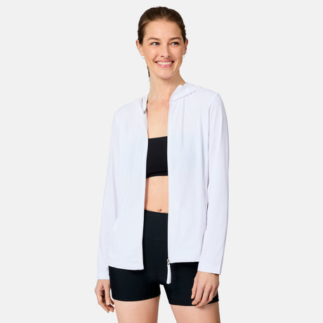 A woman smiles while wearing the Free Country Women's SunFree Full Zip UPF Sunshirt, a white hooded jacket made from recycled fabric with UPF 50+ sun protection, over a black sports bra and shorts against a plain white background.