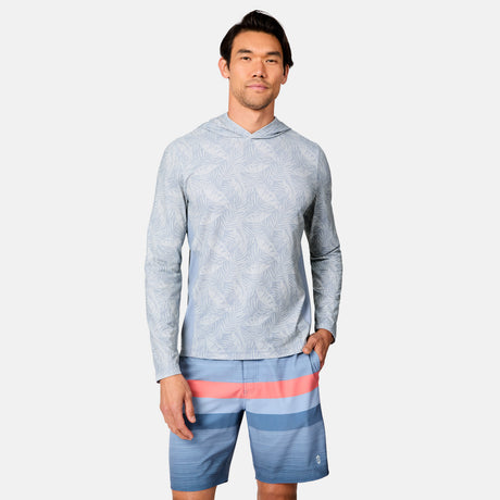 A man stands against a plain background wearing the Free Country Men's SunFree UPF Printed Hoodie in light blue with a subtle leaf pattern, and blue swim trunks with pink and red stripes, both providing UPF 50+ sun protection.