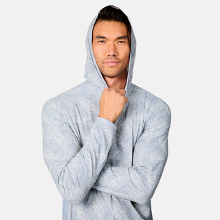 A man models the Free Country Men's SunFree UPF Printed Hoodie in light blue with a leaf pattern, standing confidently with one arm crossed and the other holding the hood near his chin against a plain background.