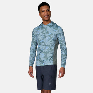 A man in a blue camouflage-patterned Men's SunFree UPF Printed Hoodie by Free Country and dark shorts stands smiling, looking to his left against a plain light gray background.