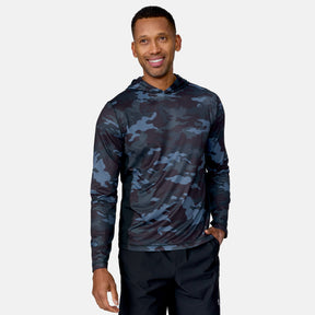 A man smiles in the Free Country Men's SunFree UPF Printed Hoodie, featuring black and dark blue camo, UPF 50+ sun protection, and black pants against a light gray background.