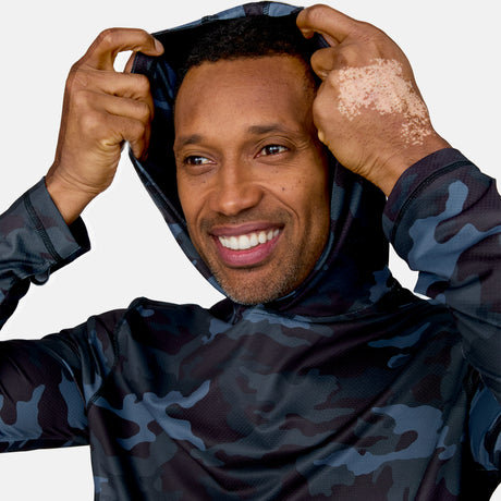 A smiling man with vitiligo on his left hand pulls the hood of a dark blue and gray camouflage Men's SunFree UPF Printed Hoodie by Free Country over his head, set against a plain white background.