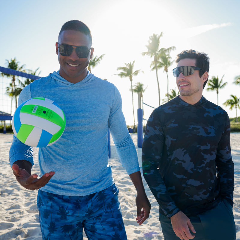 Two men in Free Country Men's SunFree UPF Printed Hoodies stroll a sandy beach with palm trees. One spins a colorful volleyball as they smile, enjoying the sun—protected by UPF 50+ for carefree outdoor fun.