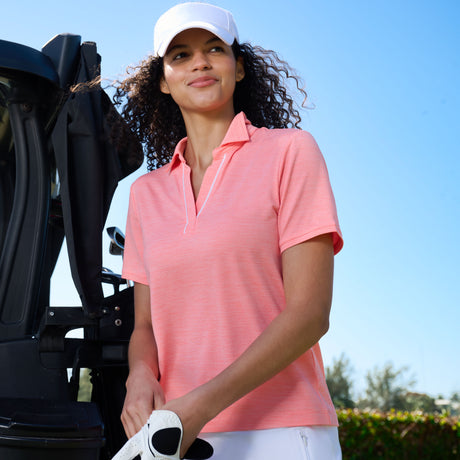 A woman in a white cap and Free Country Women’s SunFree Polo Top stands by a golf cart on a sunny day, holding a white golf glove, with blue sky and greenery in the background.
