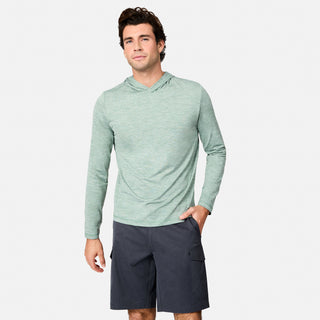 A man stands against a plain white background wearing the Free Country Men's Textured SunFree UPF Hoodie in light green and dark gray shorts with pockets, one hand in his pocket and a slight smile on his face.