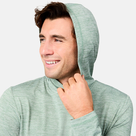 A smiling man wears the Free Country Men's Textured SunFree UPF Hoodie in light green, featuring UPF 50+ sun protection, as he looks to the side and holds his hood against a plain light background.