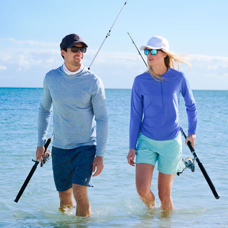 Man and woman walking in shallow water holding fishing rods on a sunny day.