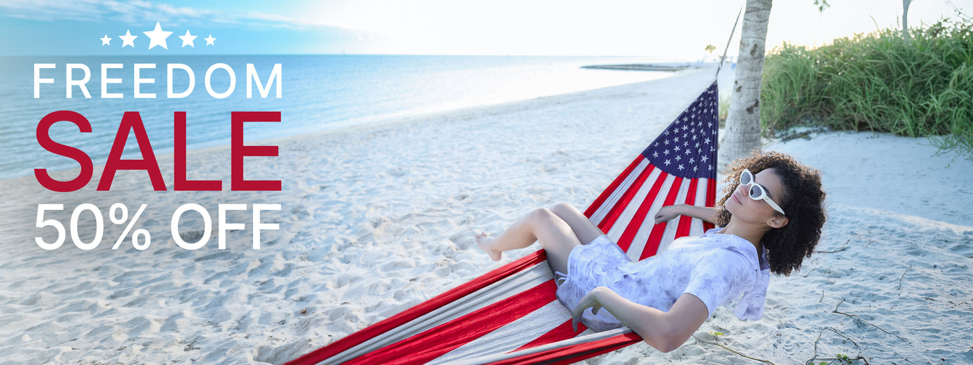 A woman relaxes in a red, white, and blue striped hammock on a sandy beach, with an American flag draped behind her. Large promotional text on the left reads 'FREEDOM SALE 50% OFF'.