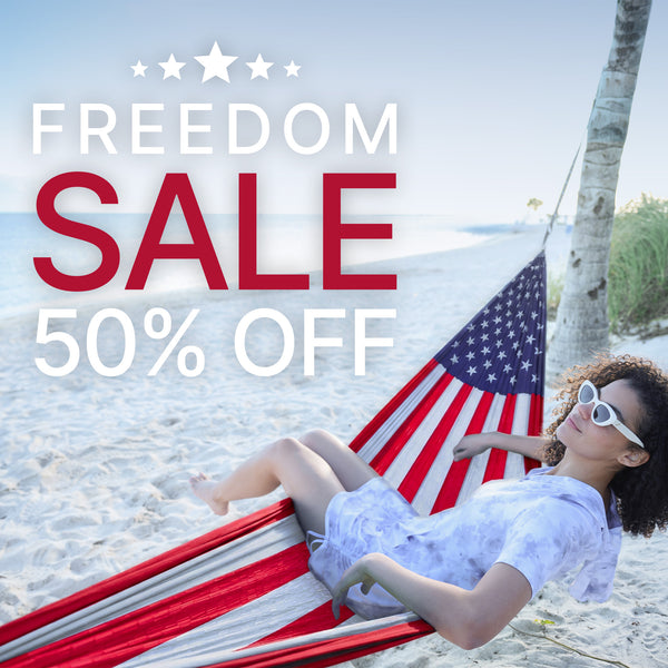 A woman wearing white sunglasses and a white dress relaxes in a red, white, and blue striped hammock on a sandy beach. Large promotional text reads 'FREEDOM SALE 50% OFF' across the top.
