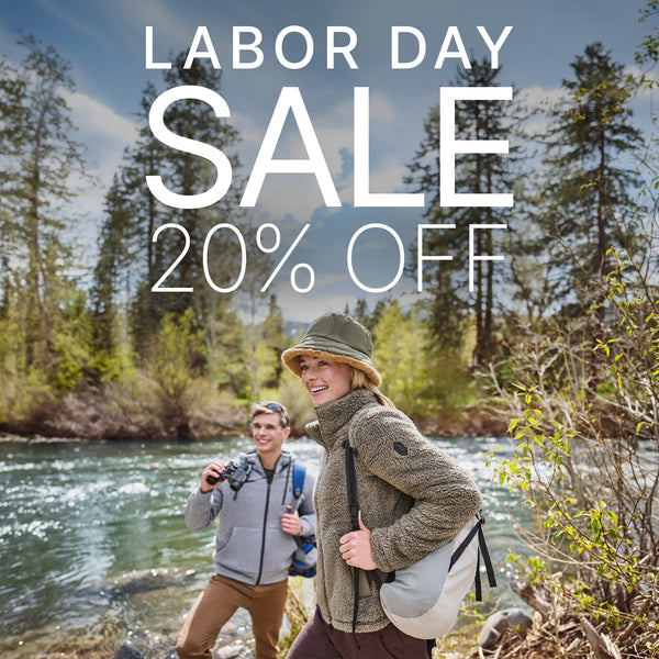 Outdoor promotional scene with two people standing by a river in a forest, overlaid with large text reading 'LABOR DAY SALE 20% OFF'. The woman wears a beige bucket hat, chunky knit jacket, and light gray shoulder bag; the man wears a gray zip jacket and brown pants.