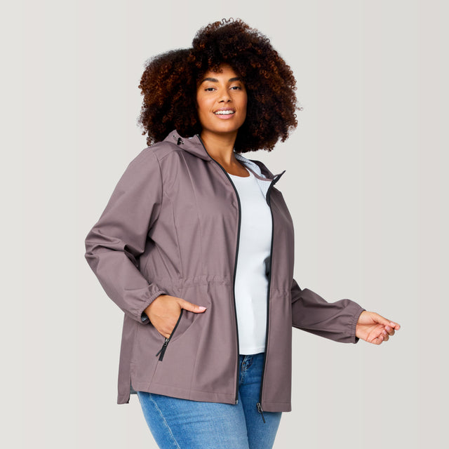 A woman with curly hair wears the Free Country Women's Plus Size X2O Anorak Rain Jacket in light brown over a white shirt and blue jeans, standing confidently with one hand in her pocket against a plain light background.