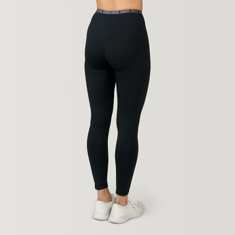 A person stands with their back to the camera against a plain background, wearing Free Country Women's Microtech® Heat Cocarber® Coffee Base Layer Bottoms in black; upper body mostly out of frame, paired with white sneakers.