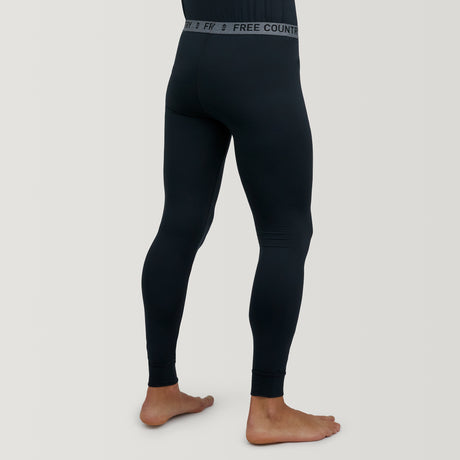 A barefoot man, shown from the waist down, wears fitted black Men's Microtech® Heat Cocarber® Coffee Base Layer Bottoms by Free Country with a visible branded waistband on a light background.