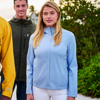 A woman wearing the Free Country Women's X2O Packable Rain Jacket in light blue stands outdoors amid greenery and sunlight. A man in a green jacket is behind her, and someone in a yellow jacket is partially visible to the left.