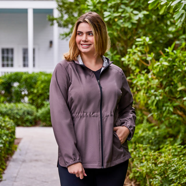 A woman with shoulder-length hair smiles outdoors on a path, wearing the Free Country Women's Plus Size X2O Anorak Rain Jacket in taupe and black pants, surrounded by lush bushes and a white building in the background.