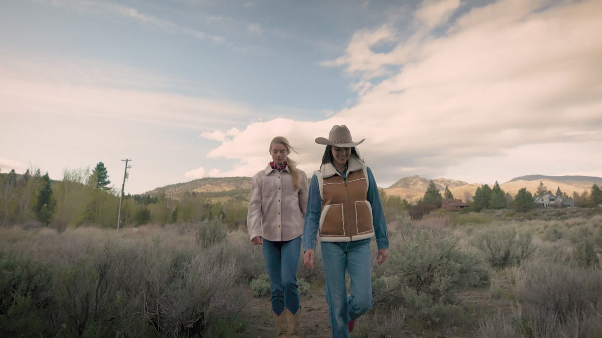 Four people walk along a dirt path through a natural landscape with shrubs, trees, and distant hills. They wear casual cool‑weather clothing and move together under a partly cloudy sky, suggesting a relaxed outdoor hike.