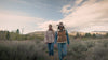 Four people walk along a dirt path through a natural landscape with shrubs, trees, and distant hills. They wear casual cool‑weather clothing and move together under a partly cloudy sky, suggesting a relaxed outdoor hike.