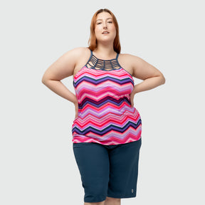 A woman models the Clearance Women's Plus Size High Neck Braided Tankini Top, featuring a pink and purple zigzag pattern and unique black strap neckline, paired with dark blue knee-length shorts against a plain light gray background.