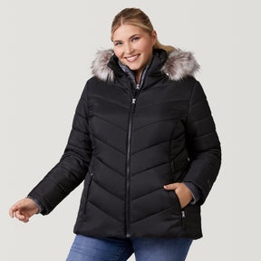 [Lola is 5'10.5" wearing a size 1X.] Woman wearing Free Country Women's Plus Size Unstoppable II Poly Air Touch Jacket in black with a full front zipper opening, attached hood with faux fur grim, patterned quilting, soft lining, inner bib, and water resistant and wind resistant stretch performance fabric. light grey background. Perfect women’s jacket for winter weather.
