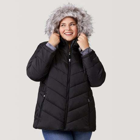 [Lola is 5'10.5" wearing a size 1X.] Woman wearing Free Country Women's Plus Size Unstoppable II Poly Air Touch Jacket in black with a full front zipper opening, attached hood with faux fur grim, patterned quilting, soft lining, inner bib, and water resistant and wind resistant stretch performance fabric. light grey background. Perfect women’s jacket for winter weather.