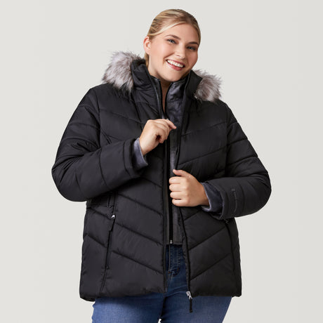 [Lola is 5'10.5" wearing a size 1X.] Woman wearing Free Country Women's Plus Size Unstoppable II Poly Air Touch Jacket in black with a full front zipper opening, attached hood with faux fur grim, patterned quilting, soft lining, inner bib, and water resistant and wind resistant stretch performance fabric. light grey background. Perfect women’s jacket for winter weather.
