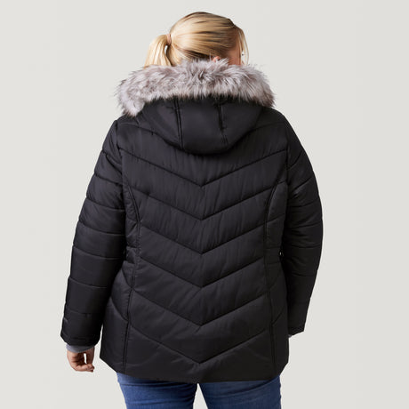 [Lola is 5'10.5" wearing a size 1X.] Woman wearing Free Country Women's Plus Size Unstoppable II Poly Air Touch Jacket in black with a full front zipper opening, attached hood with faux fur grim, patterned quilting, soft lining, inner bib, and water resistant and wind resistant stretch performance fabric. light grey background. Perfect women’s jacket for winter weather.