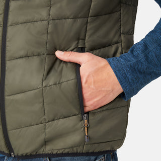 [Model is wearing a size Medium.]  Man wearing the Free Country Men's FreeCycle® Stimson Puffer Vest in green, featuring quilted design, REPREVE® poly fill, and a vertical zip, layered over a blue long-sleeve shirt with dark jeans.