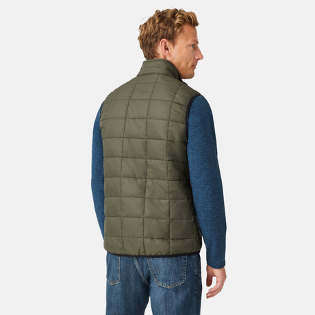 [Model is wearing a size Medium.]  Man wearing the Free Country Men's FreeCycle® Stimson Puffer Vest in green, featuring quilted design, REPREVE® poly fill, and a vertical zip, layered over a blue long-sleeve shirt with dark jeans.