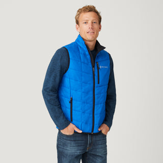 [Model is wearing a size Medium.]  Man wearing the Free Country Men's FreeCycle® Stimson Puffer Vest in blue, featuring quilted design, REPREVE® poly fill, and a vertical zip, layered over a navy long-sleeve shirt with dark jeans.