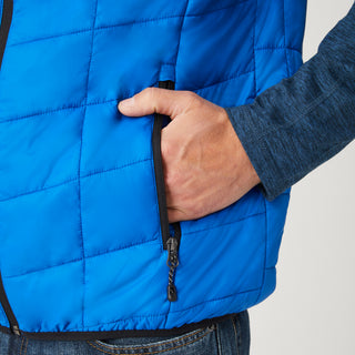 [Model is wearing a size Medium.]  Man wearing the Free Country Men's FreeCycle® Stimson Puffer Vest in blue, featuring quilted design, REPREVE® poly fill, and a vertical zip, layered over a navy long-sleeve shirt with dark jeans.