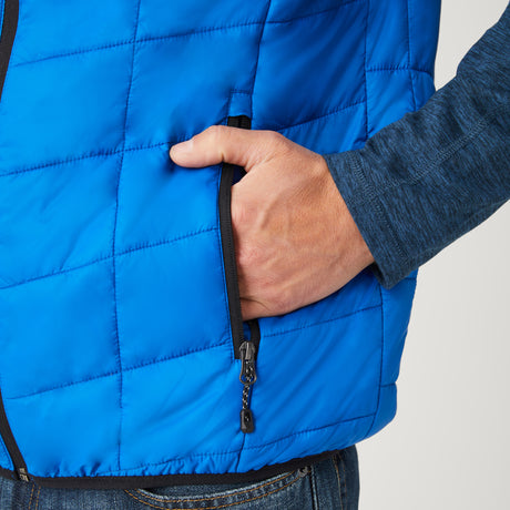 [Model is wearing a size Medium.]  Man wearing the Free Country Men's FreeCycle® Stimson Puffer Vest in blue, featuring quilted design, REPREVE® poly fill, and a vertical zip, layered over a navy long-sleeve shirt with dark jeans.