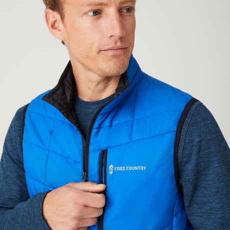[Model is wearing a size Medium.]  Man wearing the Free Country Men's FreeCycle® Stimson Puffer Vest in blue, featuring quilted design, REPREVE® poly fill, and a vertical zip, layered over a navy long-sleeve shirt with dark jeans.