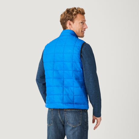 [Model is wearing a size Medium.]  Man wearing the Free Country Men's FreeCycle® Stimson Puffer Vest in blue, featuring quilted design, REPREVE® poly fill, and a vertical zip, layered over a navy long-sleeve shirt with dark jeans.